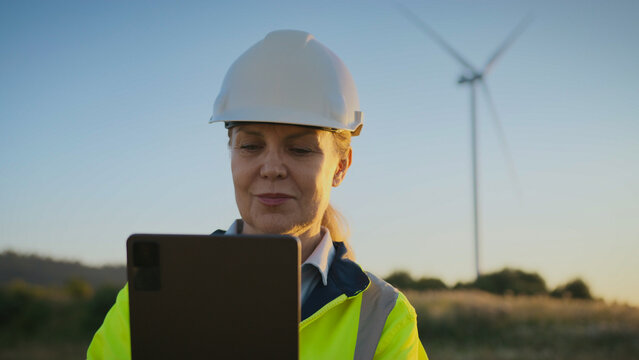 Female engineer in helmet and reflective jacket standing outdoors using digital tablet. Woman checking renewable energy data. Technician working near wind turbine in open landscape at sunset.