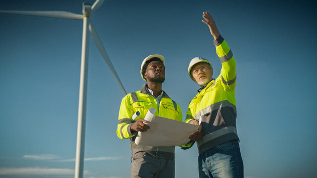 Two engineers in helmets standing near wind turbine. Workers holding blueprint. Man pointing at document while colleague observing. Team analyzing renewable energy project outdoors.