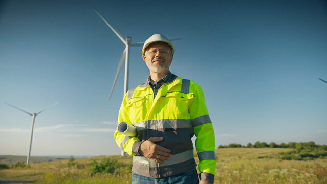 Caucasian male engineer wearing helmet and reflective jacket holding rolled blueprint standing near wind turbines. Worker looking directly at camera in open field on sunny day.