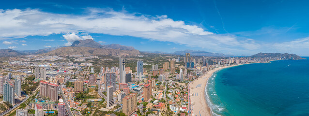 Fototapeta premium Wide panoramic view of Benidorm, Spain, showcasing the city skyline with skyscrapers, turquoise Mediterranean coastline, sandy beach, and mountain backdrop under blue sky
