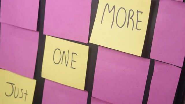 A set of yellow sticky notes standing out on a board of pink notes. With a motivating message of just one more written on them. This symbolizes extra tasks, workload, and office productivity.