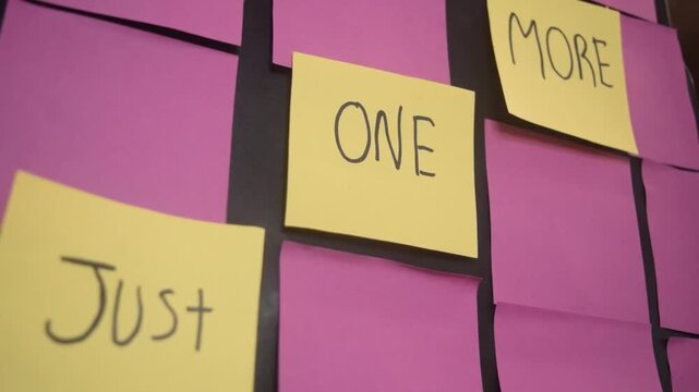 A set of yellow sticky notes standing out on a board of pink notes. With a motivating message of just one more written on them. This symbolizes extra tasks, workload, and office productivity.
