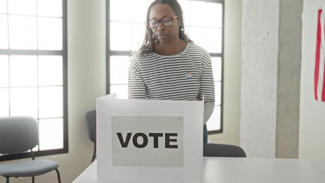 Woman in striped shirt and glasses points finger to 'i voted' sticker at a voting booth in building; civic duty pride.