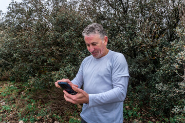 Fototapeta premium Middle-aged man in activewear smiling and scrolling his smartphone outdoors amid green foliage, enjoying nature and digital connection