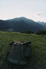 Naklejka premium Backpack resting on a grassy meadow with distant mountains and clear sky. Focus on outdoor gear, nature exploration, and rugged solitude in a calm landscape.