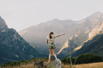 Young woman stands on a sunlit rock amid rugged mountains, reaching toward the distant valley with a sense of adventure and freedom. © SHOTPRIME STUDIO