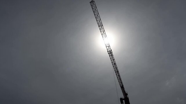 Tower crane silhouette crossing bright sun in cloudy sky creating dramatic industrial construction atmosphere with minimal composition and modern urban development mood