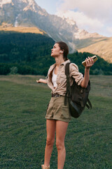 Naklejka premium Young woman with backpack standing in a lush grassy meadow, towering mountains in the distance, arms raised, holding a book, conveying adventure, outdoor lifestyle, exploration, and fresh air.