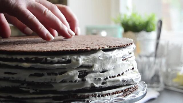 Close-up of female hands spreading smooth creamy frosting on chocolate sponge cake layers while preparing a homemade layered dessert.