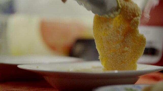 Close view of raw chicken coated in breadcrumbs being dipped into egg mixture with kitchen tools and ingredients blurred in background, highlighting texture and preparation steps