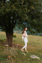 Naklejka premium Relaxed woman strolling through a sunlit countryside, dressed in white, enjoying fresh air and nature, a casual lifestyle moment by a large tree.