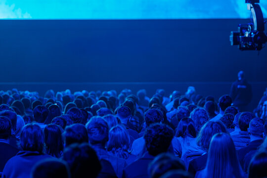 Large audience seated in blue lit conference hall, watching presentation on stage. Moody lighting, crowded rows, live event atmosphere and focused attention.