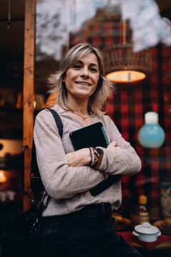 Smiling mature woman standing outdoors near cafe window holding digital tablet, happy female entrepreneur enjoying business success in city, authentic lifestyle of successful professional.