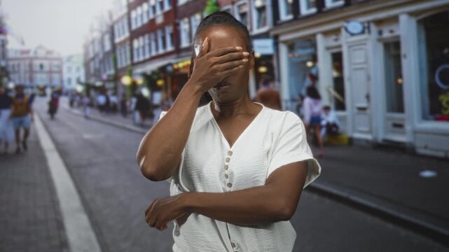 Woman with hand covering eyes on a busy city street near storefronts, visible forearm and closed eyes gesture; frustration reflection.