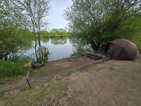 Professional fishing camp on a peaceful riverbank with a camouflage bivvy tent, rods, and equipment. Carp fishing setup in a scenic natural landscape.