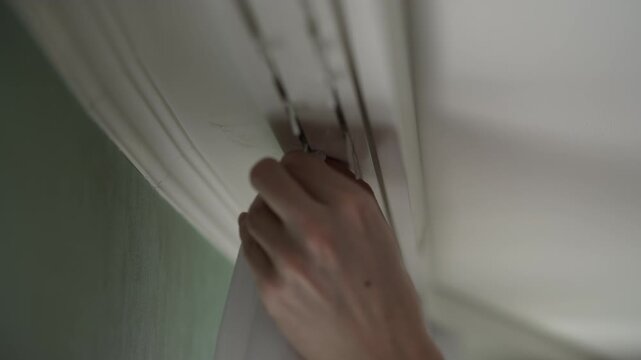 Detailed view of person hands attaching curtain hooks to gliders of ceiling mounted track, methodically completing installation process in home interior. Shooting in slow motion.