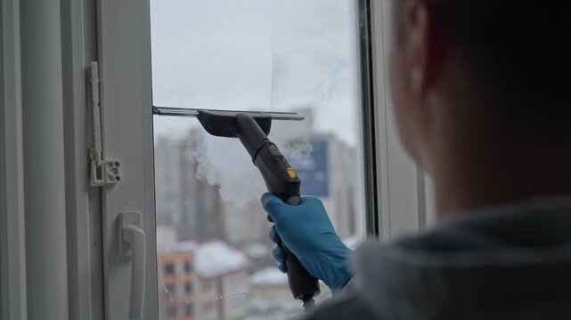 Rear view of professional cleaning service employee washing dirty window with special steam cleaner. Homemaker in rubber gloves removes dirt from glass surface inside modern apartment, slow motion.