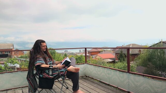Authentic Bohemian Man with Long Loose Hair wearing Colorful Baja Hoodie and Harem Pants Reading a Paperback Book Turning Pages and Swinging Crossed Leg on Scenic Terrace with Ocean View Backdrop