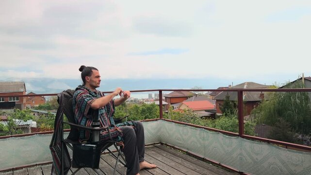 Bohemian Caucasian Man with Hair Bun Wearing Ethnic Colorful Poncho Stretching Arms Up and Resting in Camping Chair on Scenic Terrace with Sea and Mountain View after Deep Thinking Authentic Shot