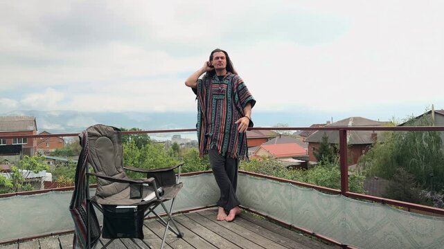 Bohemian Traveler with Long Flowing Hair Standing on Veranda Gathering Hair then Jumping to Sit on Balcony Railing Overlooking Scenic Sea Harbor and Mountains at Cloudy Spring Day Authentic Shot