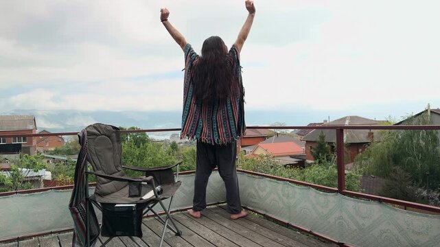 Free-Spirited Bohemian Man with Long Loose Hair wearing Ethnic Serape Standing on Terrace Stretching Arms Up and Leaning on Railing with Crossed Legs overlooking Coastal Bay View Shot