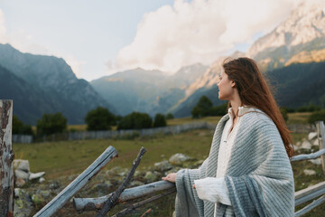 Naklejka premium Young woman with long hair gazes at alpine mountains, wrapped in a cozy shawl, standing beside a weathered wooden fence, enjoying a calm lifestyle moment in nature