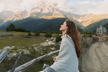 Fototapeta premium A woman stands along a country road wrapped in a blanket, gazing at distant mountains under a clear sky, conveying calm, solo travel and reflective lifestyle.