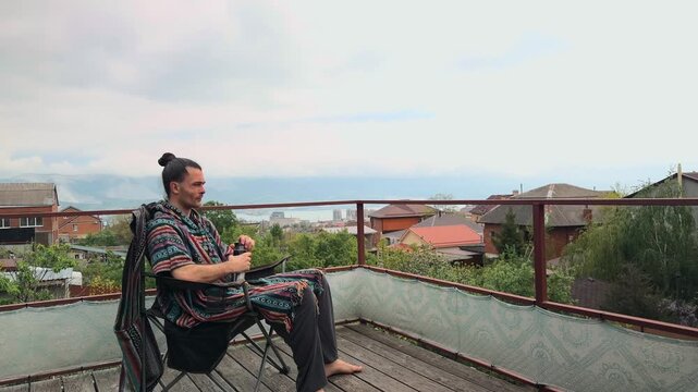 Relaxed Adult Bearded Man with Hair Bun Wearing Ethnic Boho Poncho and Harem Pants Sitting in Camping Chair on Terrace and Drinking from Thermos Tumbler at Cloudy Day with Sea Bay View Profile Shot