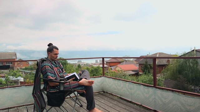 Relaxed Caucasian Man with Hair Bun Wearing Ethnic Colorful Poncho and Grey Harem Pants Sitting in Chair on Wooden Terrace with Sea View Taking Out and Reading a Book on a Cloudy Warm Day Shot