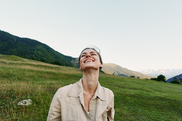 Naklejka premium Cheerful woman outdoors, smiling up at the sky in a sunlit meadow, relaxed posture, natural colors, wide landscape, mountains in distance, lifestyle moment