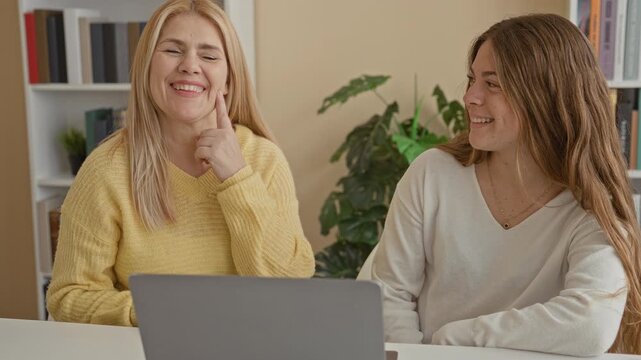 Woman and daughter lean over a laptop, daughter kisses mother's cheek while mother laughs with hand near chin in a livingroom study; family bonding joyful.