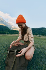 Naklejka premium Woman in an orange beanie sits on grass beside a backpack in a mountain meadow, enjoying outdoor life and natural scenery and sunlight warms her face under a clear blue sky