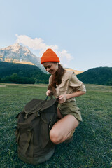 Naklejka premium Young woman outdoors kneels beside a large backpack in a green meadow with distant mountains, smiling softly as she begins unpacking gear for a day in nature.