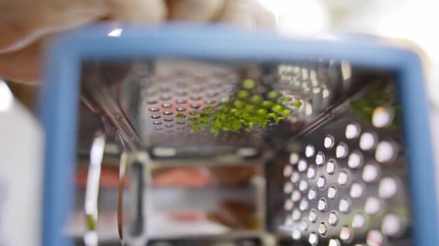 Close view of lemon zest being grated on metal grater, fine citrus peel falling through holes, food preparation step adding flavor for cooking in simple kitchen setup