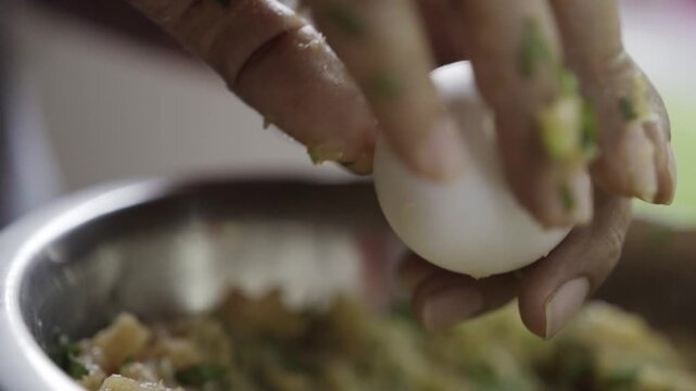 Close view of hand cracking egg into chicken mixture in metal bowl, liquid egg falling into ingredients, step of meatball preparation in simple homemade kitchen process