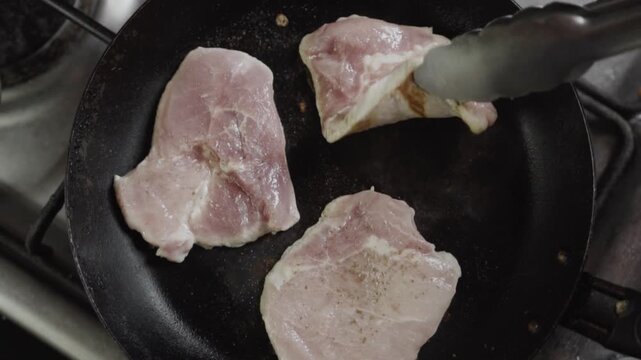 High angle close up of raw pork loin steaks being flipped with metal tongs in a hot pan, showing browning and searing process.