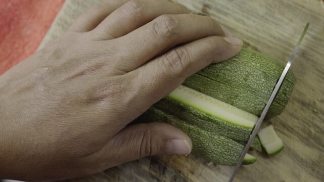 Closeup view of hand cutting fresh zucchini into slices on wooden board, simple kitchen scene highlighting ingredient preparation and healthy cooking process