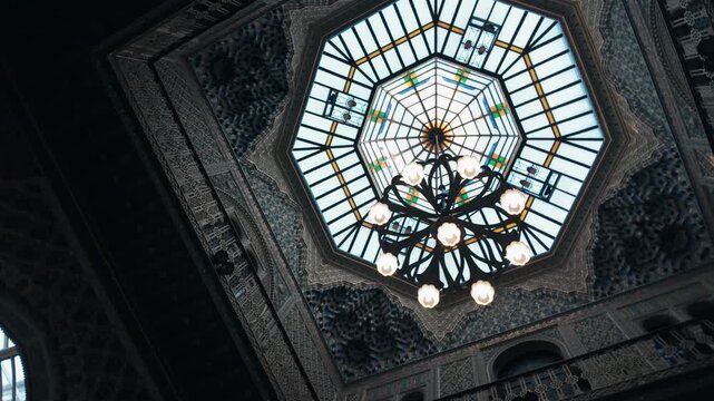 Low angle view of a majestic octagonal glass skylight featuring intricate floral stained glass patterns and a classic wrought iron chandelier hanging in a historic luxury palace or grand hall