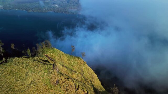 Low clouds drifting over volcanic mountain ridge above deep blue crater lake, cinematic aerial scene with steep grassy slopes and dramatic morning light aerial tilt-up drone footage
