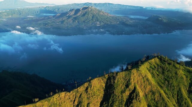 Volcano caldera lake in Indonesia with sunlit mountain slopes and clouds, Mount Batur visible in the background, aerial drone footage