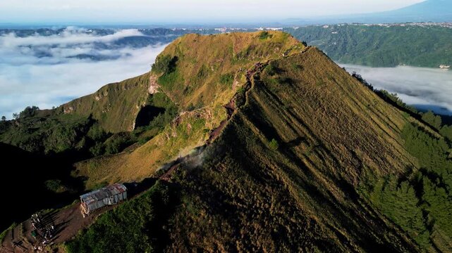 Hikers trekking along volcanic ridge trail across Mount Batur crater rim with rugged terrain and steep slopes as volcanic smoke rises in soft morning light smooth aerial drone footage