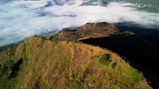People walking along narrow mountain trail above sea of clouds in early morning light across Mount Batur volcanic crater rim with steep green slopes and dramatic terrain aerial arc drone footage