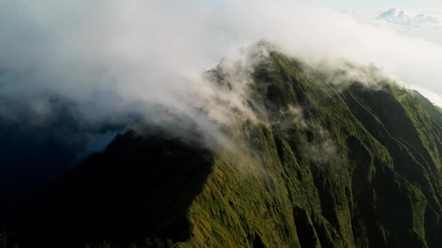 Clouds moving across volcanic mountain ridge with steep green terrain and dramatic sunlight in Indonesia, drifting fog revealing textured slopes and rugged mountain relief aerial drone footage