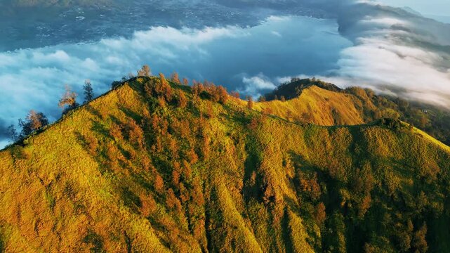 High altitude mountain ridge with steep slopes opening to Mount Batur caldera and crater lake in golden hour light across Bali landscape cinematic aerial drone footage