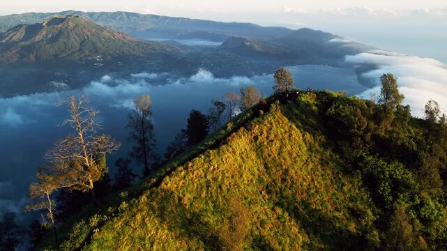Hikers on mountain ridge overlooking volcanic caldera and crater lake on Trunyan Hill with soft clouds drifting over mountain ridge in golden hour light across high altitude terrain aerial drone view