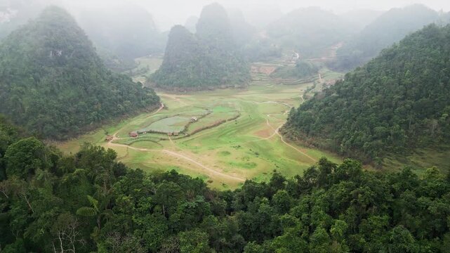 Rainy mountain valley in Northern Vietnam with low clouds drifting through forested hills and karst formations over agricultural fields creating dramatic moody atmosphere aerial drone view