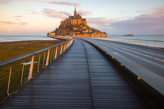 View of the historic Mont Saint-Michel abbey at sunset with the long wooden bridge leading towards the island under a soft colorful sky Mont Saint-Michel, Normandy, France.