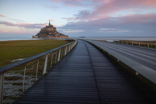 View of the historic Mont Saint-Michel abbey at the end of a long wooden pedestrian bridge under a soft pink sunset sky Mont Saint-Michel, Normandy, France.