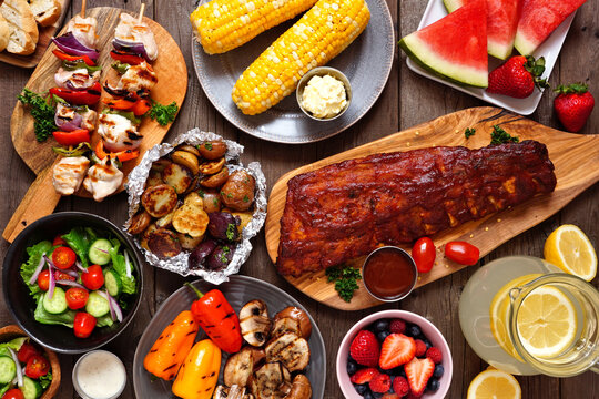Summer BBQ or picnic food concept. Assortment of grilled meats, vegetables, fruits, salad and potatoes. Top down view table scene over a dark wood background.
