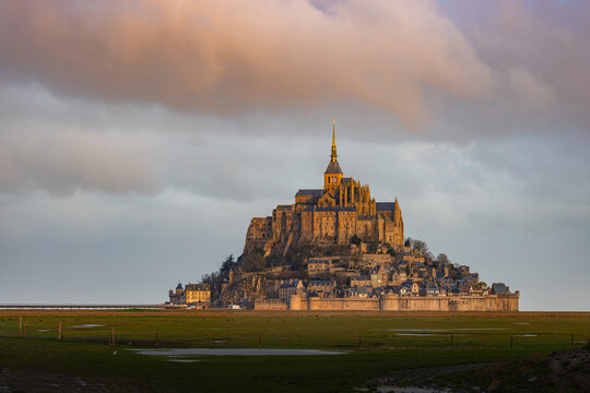View of Mont Saint-Michel abbey illuminated by golden sunset light under dramatic clouds with green salt marshes in the foreground Mont Saint-Michel, Normandy, France.